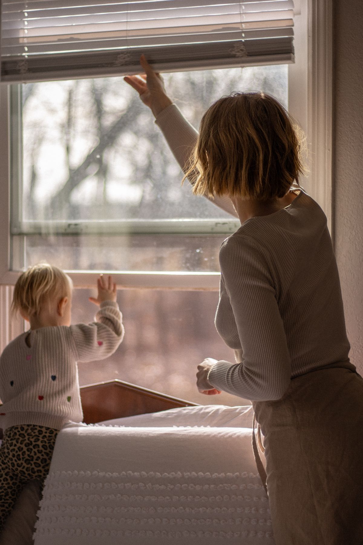 A parent lifting a blind as a child looks towards the window